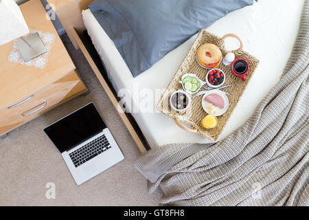 High Angle View of Open Laptop on Floor Beside Unmade Bed in Hotel Room with Wicker Breakfast Tray Stock Photo