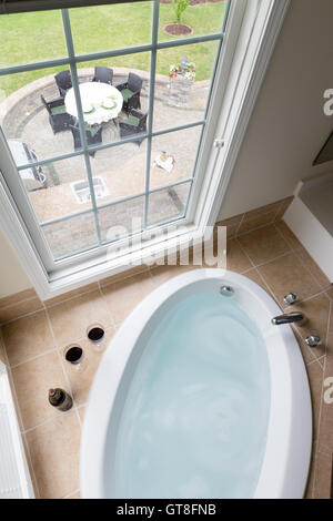 Modern sunken oval bathtub full of clean water in a brown tile surround overlooking a brick patio with outdoor dining through a Stock Photo