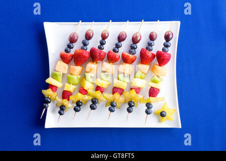 Healthy fruit kebabs with tropical seasonal summer fruit arranged diagonally on a modern rectangular plate over a texture blue b Stock Photo