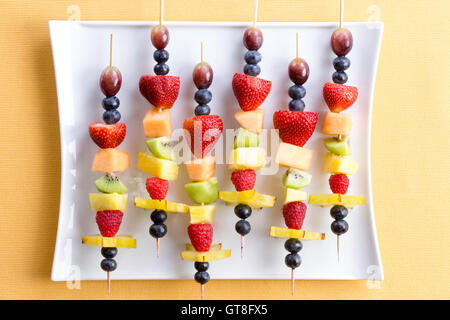 Healthy tropical summer fruit kebabs in a colorful arrangement on a modern white rectangular plate on a textured yellow table, o Stock Photo