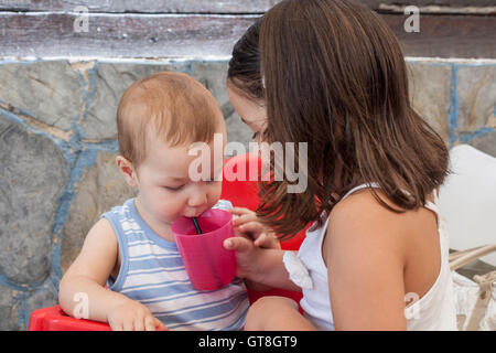 Little sister helping to her baby brother to drink juice with straw. She is holding the plastic glass Stock Photo