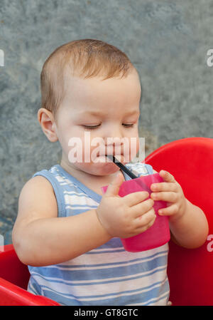 Baby boy drinking by himself with straw. He is seated in high chair at terrace restaurant Stock Photo