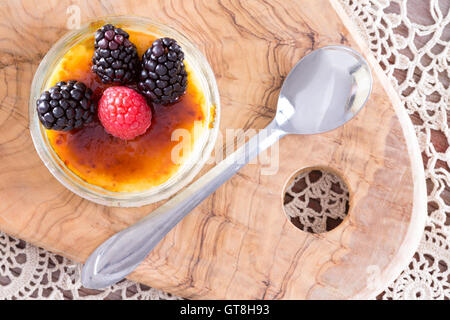 Scrumptious creme brulee topped with red and black raspberries from point of view angle with spoon over doily and wooden tray Stock Photo