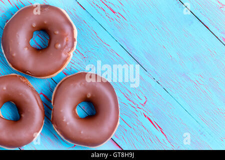 Three crispy cream filled chocolate ring donuts topped with chocolate icing on a rustic turquoise blue wooden background with co Stock Photo