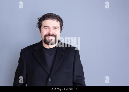 Single smiling handsome dark haired and bearded middle aged man wearing black shirt and blazer over gray background with copy sp Stock Photo