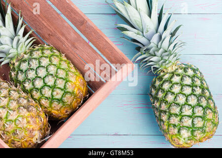 Three fresh pineapples, two in a wooden crate and one alongside on a blue wooden table lying on their sides viewed close up from Stock Photo
