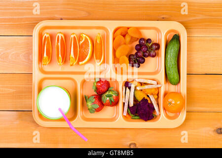 Lunchtime plastic tray filled with orange wedges, dried apricot, grapes, pickle, strawberry and other ingredients next to cup of Stock Photo