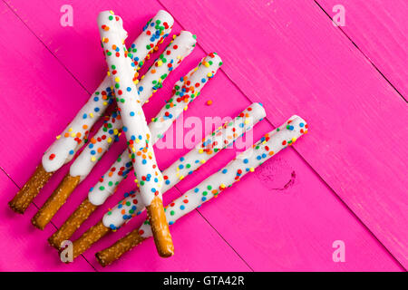 Six long pretzel sticks coated with white chocolate and various colored candy sprinkles over bright pink wooden background Stock Photo
