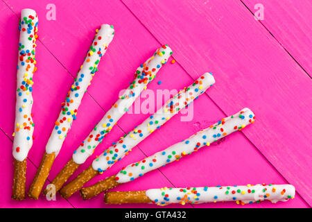 Six long pretzel sticks coated with white chocolate and various colored candy sprinkles arranged in a ray shape over bright pink Stock Photo