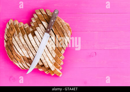 Top down view of freshly baked and sliced heart shaped bread with sharp knife over it on pink wooden table background with copy Stock Photo