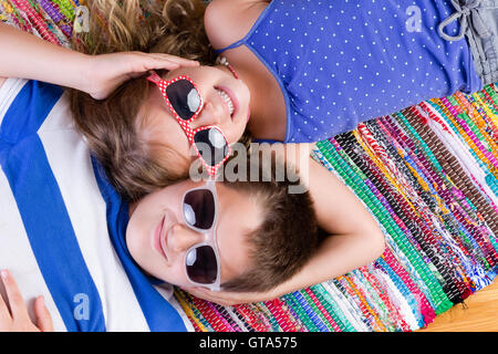 Cute brother and sister in sunglasses laying down on colorful rug while holding each others heads Stock Photo