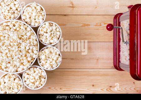 Dishes of fresh popcorn arranged in a decorative circle around a larger center bowl alongside a machine on a wooden table with c Stock Photo