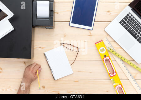 Man at work on a retail carpenters desk with a cash register, tablet, laptop, builders level and ruler and a blank notepad with Stock Photo