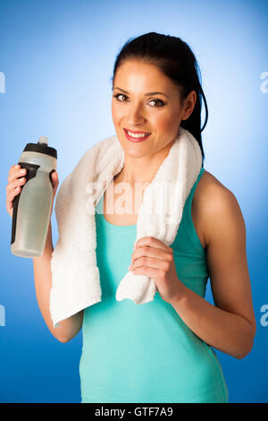 Woman rests after fitness workout with towel around her neck drinking water over blue background Stock Photo