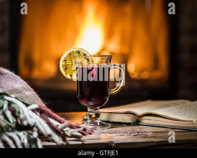 Hot mulled wine and a book on the wooden table. Fireplace with warm fire on the background. Stock Photo