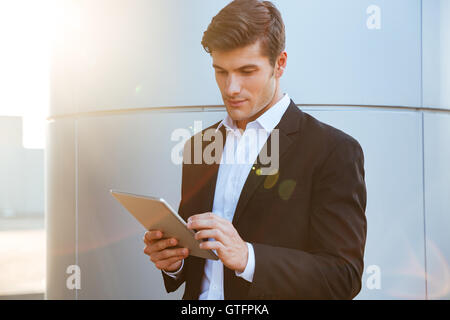 Attractive young serious businessman standing outdoors and using pc tablet Stock Photo