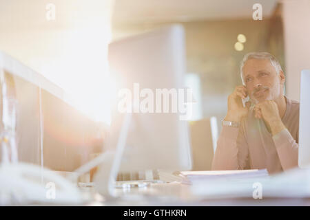 Businessman talking on cell phone at computer in sunny office Stock Photo