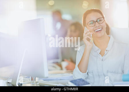 Smiling businesswoman talking on cell phone at computer in office Stock Photo
