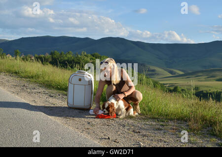 Woman taking care of her dog giving her water from a dog's water container on a mountain road on sunny day Stock Photo