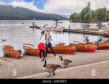 Two Asian girls taking photos with a selfie stick on the shores of Lake Windermere, Cumbria, England, UK Stock Photo