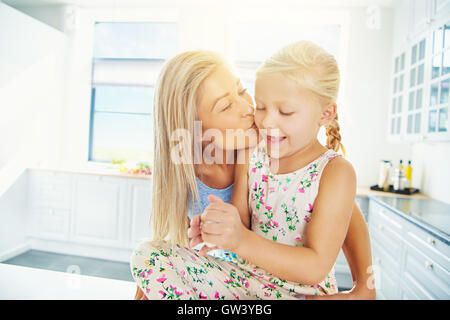 Beautiful young blond mother kissing daughter in ponytails and dress while she plays with bread dough Stock Photo