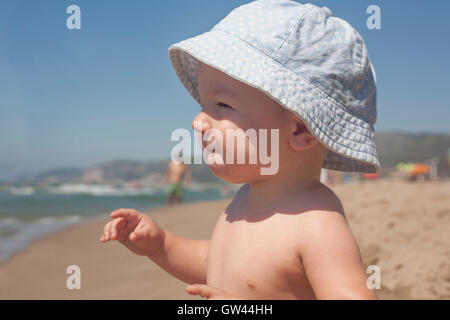 Happy baby boy with blue hat sitting on the sand and observing the seashore on the beach Stock Photo