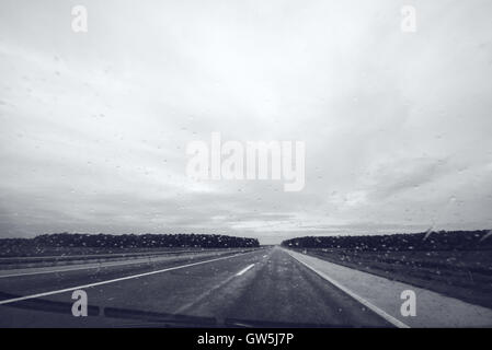 Raindrops on car windshield glass, monochromatic image of open highway through vehicle windscreen on a rainy day Stock Photo