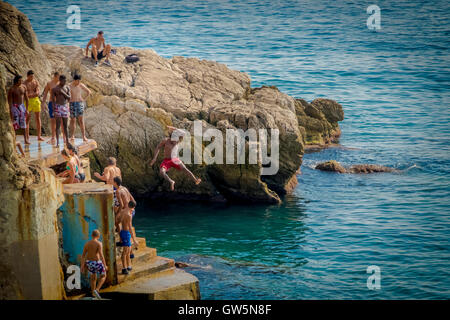 Young boys dive off a platform into the blue waters of the Mediterranean Sea while others watch and wait their turn Stock Photo