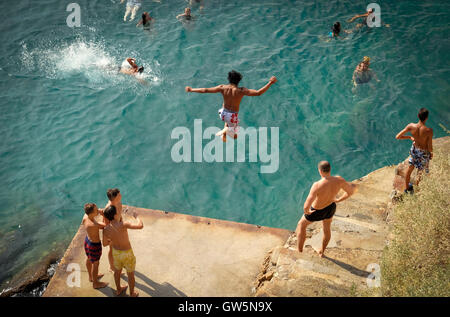 Young boys dive off a platform into the blue waters of the Mediterranean Sea while swimmers watch below Stock Photo