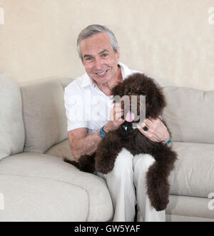 Clean cut slim and grey senior elderly man sits on a beige couch, with his pet companion labradoodle  puppy dog on his lap. Stock Photo