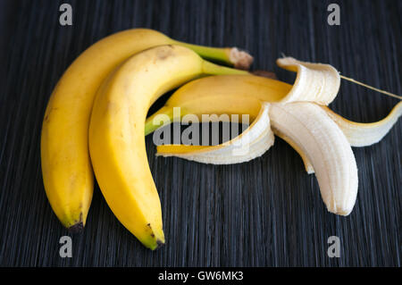 three ripe bananas, one peeled half on a dark background close-up Stock Photo