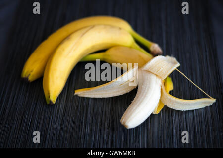three ripe bananas, one peeled half on a dark background close-up Stock Photo