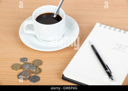 Cup of black coffee with a spoon in it on a wooden table with notepad and coinage Stock Photo