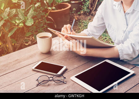 Woman hand writing notebook and phone, tablet on table in garden at coffee shop with vintage toned. Stock Photo