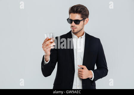 Focused young businessman in suite and sunglasses looking at whiskey in glass over gray background Stock Photo