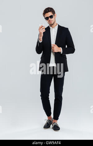 Full length portrait of a smiling happy businessman in suit holding glass of whiskey over gray background Stock Photo