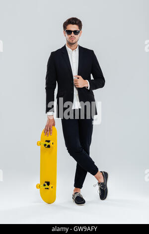 Serious young businessman in sunglaasses and suit leaning on skateboard with legs crossed over gray background Stock Photo