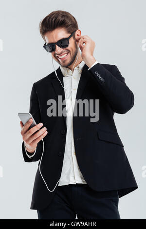 Smiling handsome young businessman listening music with earphones holding smartphone over gray background Stock Photo