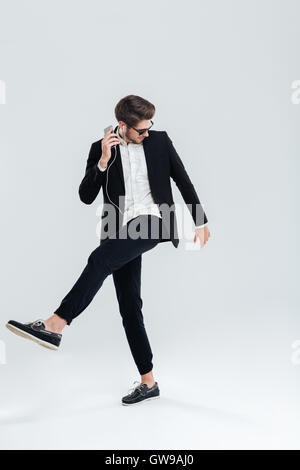 Happy attrative young businessman in black suit listening music with earphones and dancing over gray background Stock Photo