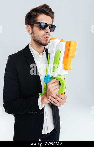 Handsome businessman in black suglasses holding colorful water gun over gray background Stock Photo