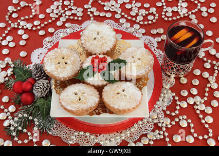 Christmas mince pie cakes with mulled wine, holly, baubles, winter greenery and silver decorations on red background. Stock Photo