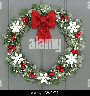 Christmas wreath with red baubles and bow and white snowflake decorations and snow covered blue spruce fir over grey front door Stock Photo