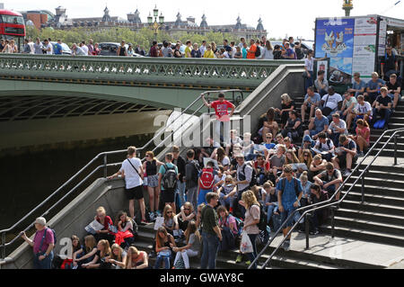 Crowds of young tourists and visitors sit on the steps of Westminster Bridge, London, next to Big Ben and River Thames Stock Photo