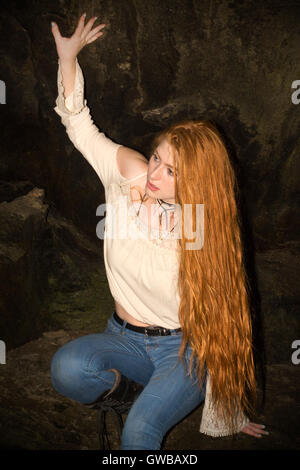 Beautiful red head in skinny jeans, white top and black boots, looking trapped or lost in a small cave at an old train tunnel. Stock Photo