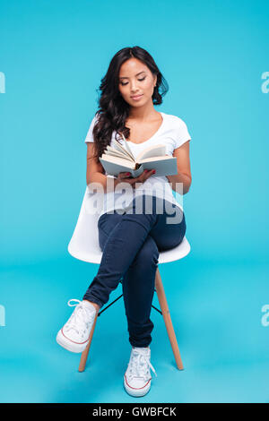 Pensive young attractive vietnamese girl reading book and sitting on chair isolated on the blue background Stock Photo