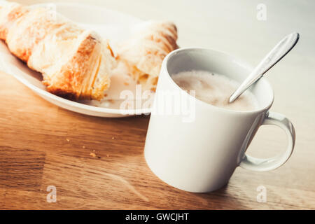 Cappuccino and croissant. White cup of coffee with milk foam stands on wooden table, closeup photo with soft selective focus. Vi Stock Photo