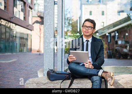 Young  asian business man holding a digital tablet sitting outside on city street looking thoughtful and concerned Stock Photo