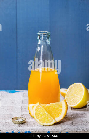 Opened glass bottle of citrus orange and lemon lemonade, standing with sliced fruits on stone board with blue background. With c Stock Photo