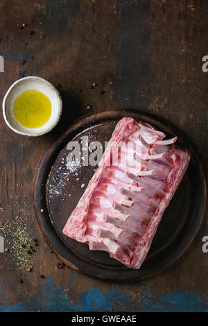 Raw uncooked rack of lamb on wood chopping board with salt, pepper, dry herbs and olive oil over old wooden background. Top view Stock Photo