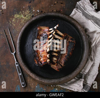 Whole grilled bbq rack of lamb, served with seasoning,  meat fork and textile napkin on clay tray over old wooden background. To Stock Photo